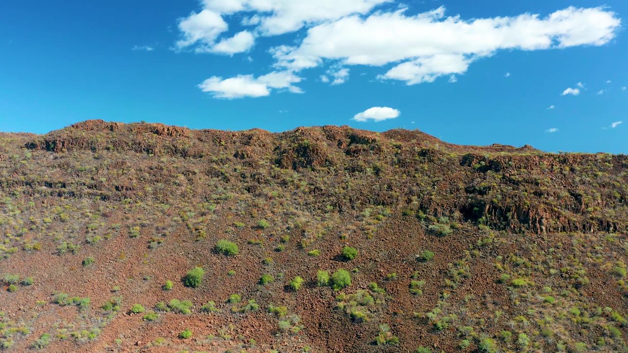 Third frame in San Javier, Baja California Sur, with a wide view of reddish rocky terrain, scattered shrubs, a ridgeline, and no visible people.