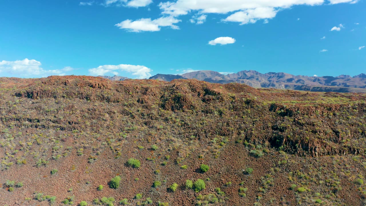 Opening frame over reddish rocky terrain near San Javier, Baja California Sur, with scattered green shrubs, distant mountains, blue sky, small white clouds, and no visible people.