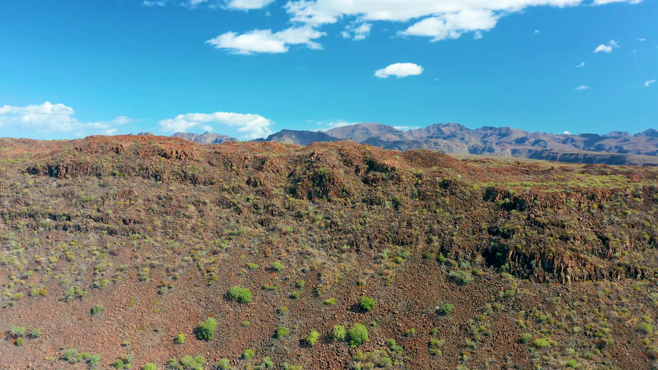 Early frame showing the same San Javier rocky slope from above, with a dark ridge line across the midground, green shrubs on red soil, mountains on the horizon, and no visible people.