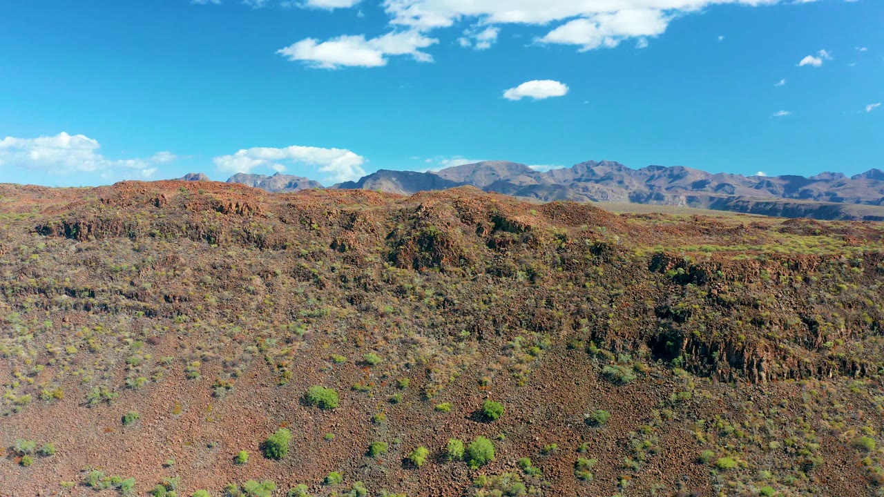 Frame at two seconds with red-brown San Javier terrain filling the lower view, sparse shrubs dotted across the slope, a broken rocky ridge, distant mountain range, and no visible people.