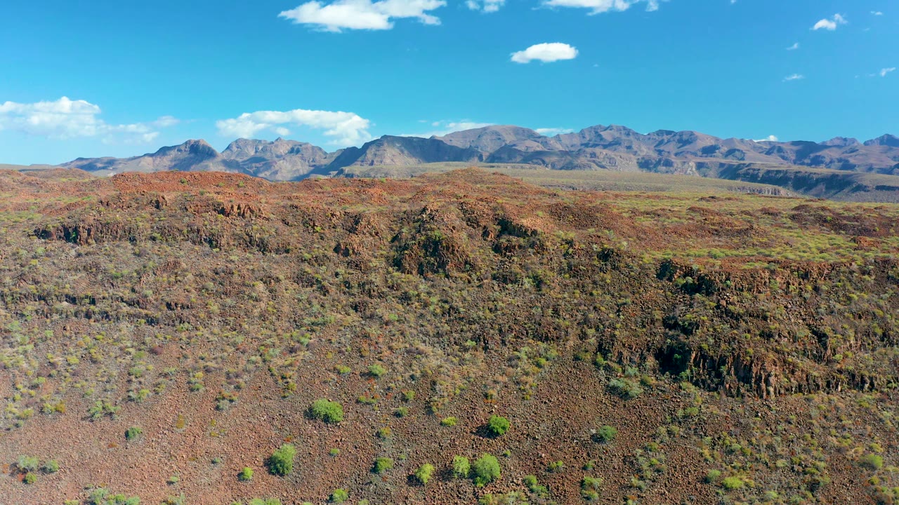 Reddish desert slope and plateau with shrubs, mountains, and scattered clouds.