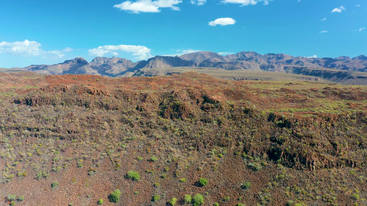 Opening view of red rocky desert terrain near San Javier, Baja California Sur, with scattered green shrubs, distant mountains, small white clouds, and no visible people.