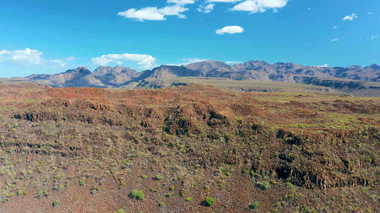 Mid-clip frame shows wide red-brown desert foreground near San Javier, Baja California Sur, with sparse shrubs, uneven rocky ridges, distant mountains, and no visible people.