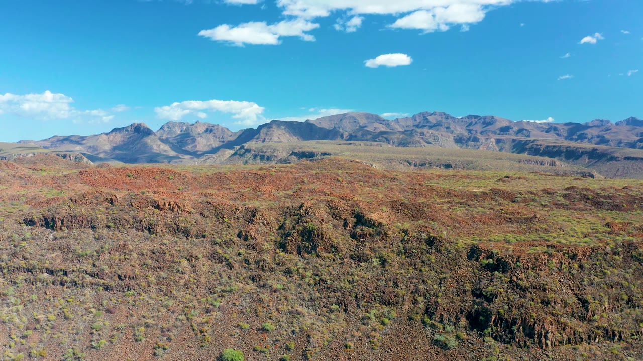 Opening frame of San Javier, Baja California Sur, with red-brown rocky terrain in the foreground, distant mountains on the horizon, scattered clouds, and no visible people.