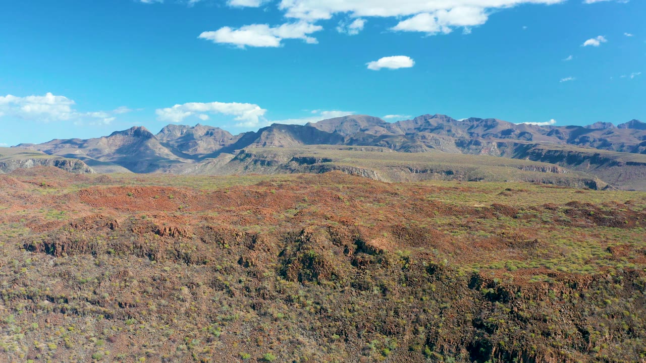 Opening frame of San Javier, Baja California Sur, showing reddish plateau terrain, sparse vegetation, distant mountain ranges, small white clouds, and no visible people.
