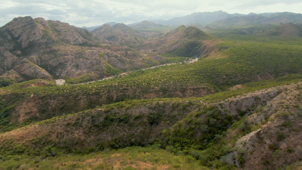 Aerial view over Cabo Pulmo, Baja California Sur, Mexico, showing rocky arid mountains, green vegetation, a pale winding road in the midground, distant hazy ridges, and no visible people.