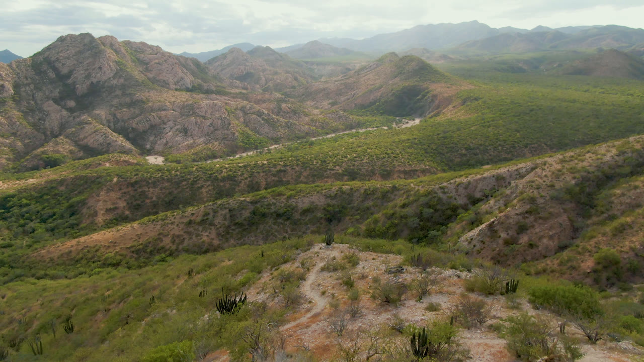 Aerial view of rocky mountains, green scrub, and a pale winding road under clouds.