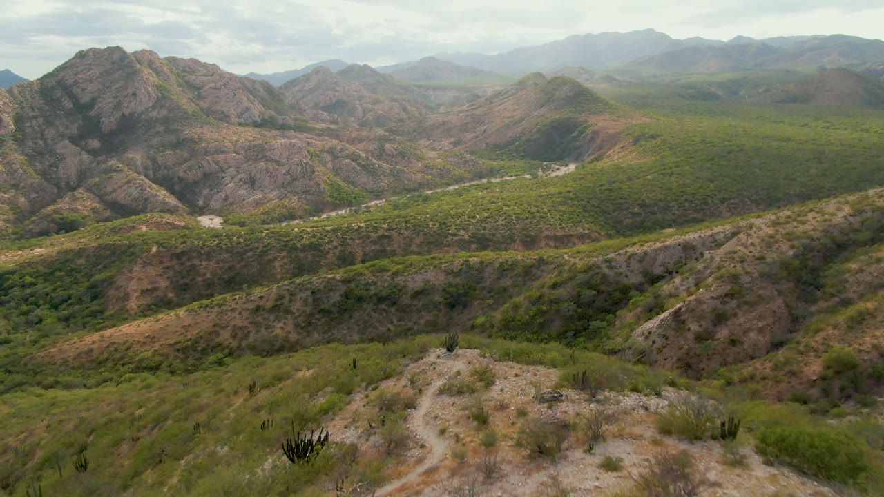 Second frame in Cabo Pulmo shows the pale road cutting across the green-brown valley below rocky slopes, with hazy mountains in the distance and no visible people.