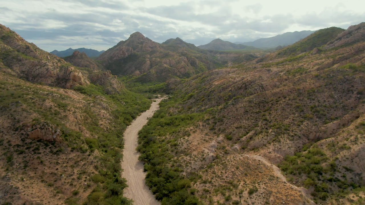Aerial view in Cabo Pulmo with a rocky ridge filling the right side, a pale winding road or wash on the left, layered mountains beyond, and no visible people.