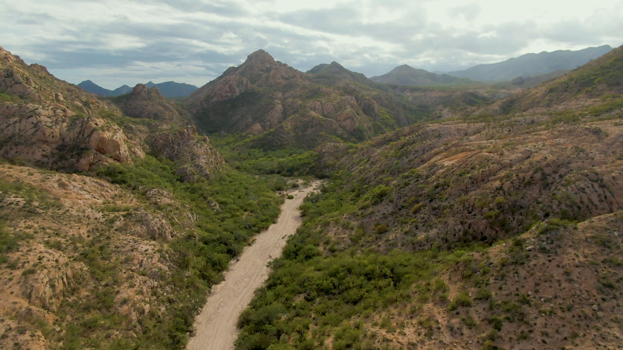 Aerial frame from Cabo Pulmo, Baja California Sur, Mexico, showing the dirt road nearer the lower center, green valley vegetation, tan rock ridges, layered mountain peaks, cloud-filtered daylight, and no visible people.