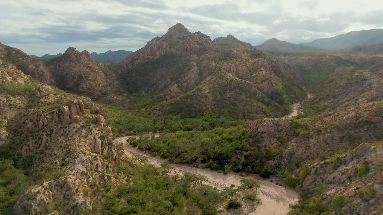 Aerial frame in Cabo Pulmo showing the dirt road curving through the valley floor, with jagged brown hills, green brush, hazy mountains, cloudy daylight, and no visible people.