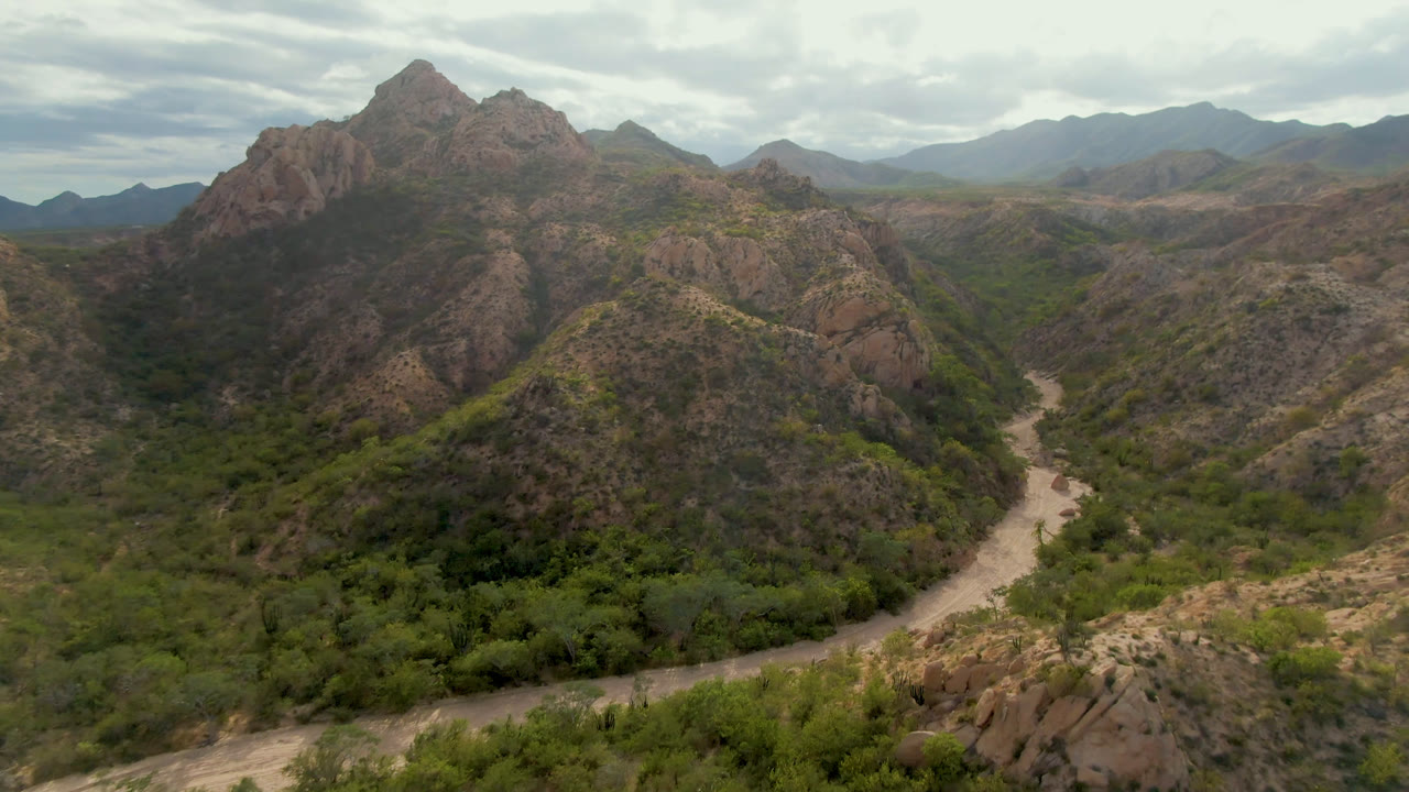 High aerial composition over Cabo Pulmo terrain, showing the dirt road lower in frame, scrub-covered slopes, rocky peaks, distant mountains, and no visible people.