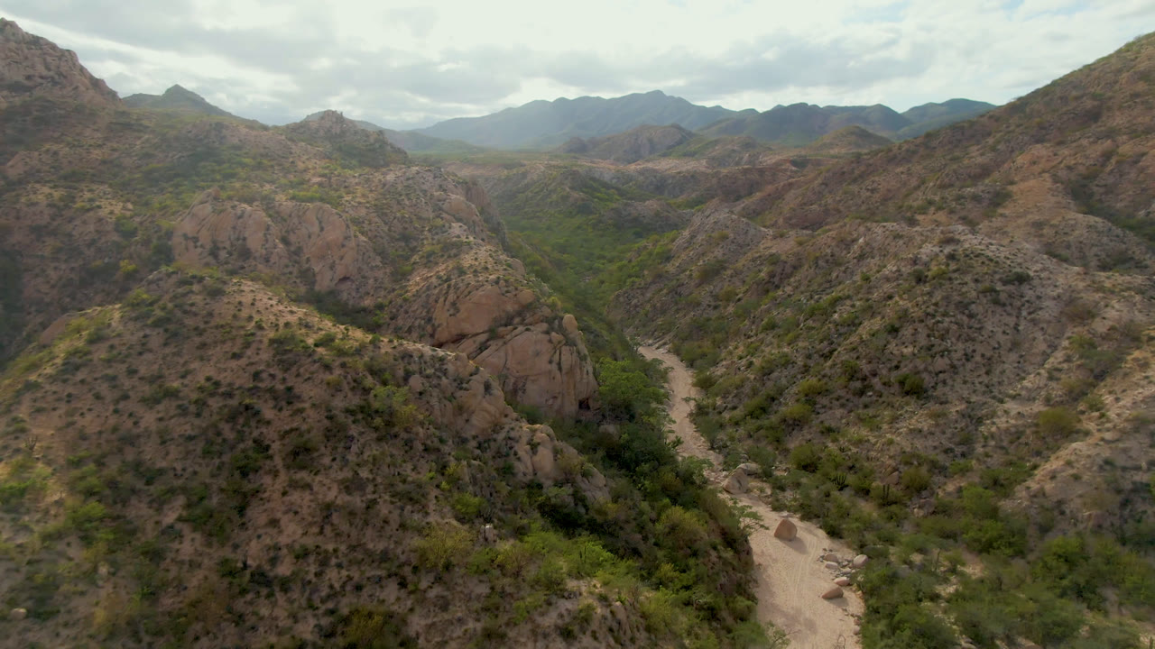 Cabo Pulmo aerial frame showing the dirt road slightly deeper between scrub-covered hills, reddish rock faces, distant ridges, and no visible people.
