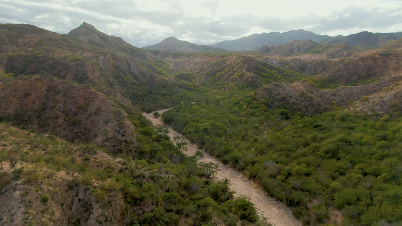 Aerial frame from Cabo Pulmo, Baja California Sur, showing a pale dirt road entering a green valley below brown rocky hills, with distant mountains and no visible people.