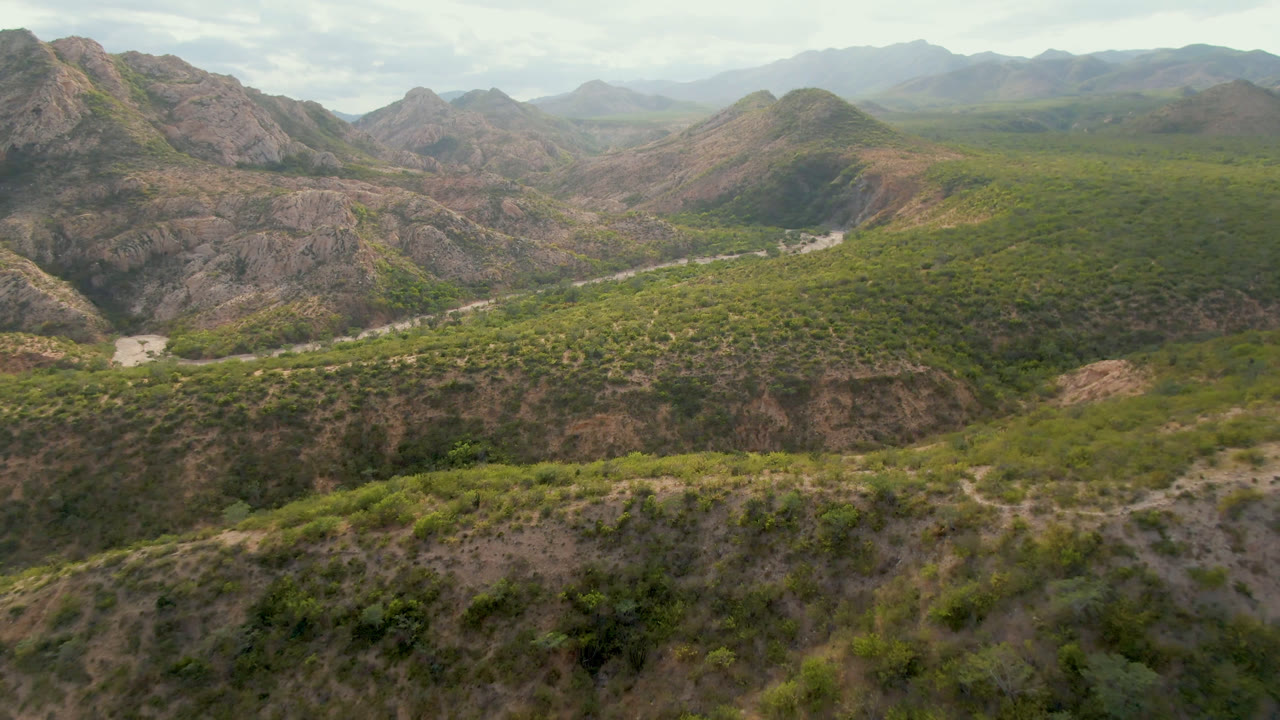 Aerial frame from Cabo Pulmo, Baja California Sur, showing rocky foreground hills, a green valley, distant mountain ridges, hazy daylight, and no visible people.