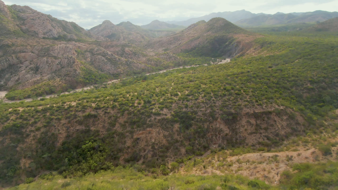 Aerial view over Cabo Pulmo, Baja California Sur, showing a green desert valley, rocky hills, distant mountains, and no visible people at the start of the clip.