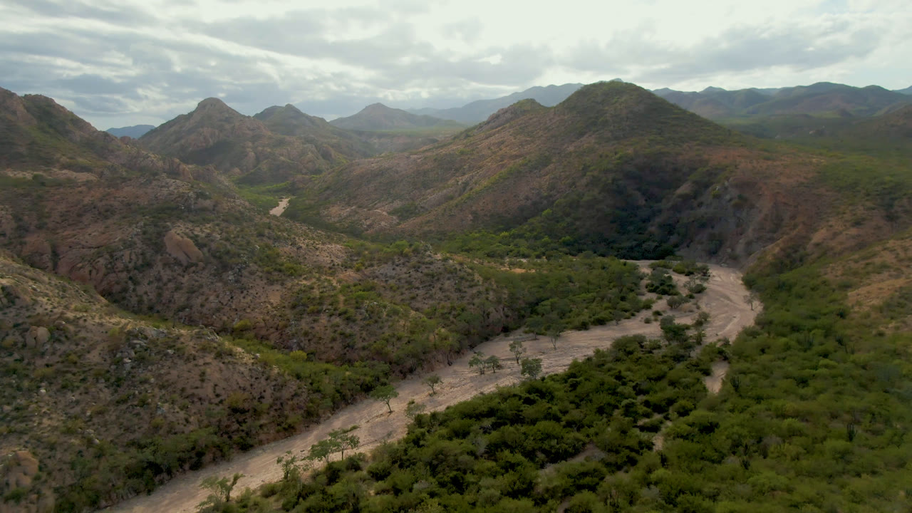 Aerial view in Cabo Pulmo, Baja California Sur, with the pale valley road curving from the lower frame toward scrub-covered ridges, distant mountains, cloud cover, and no visible people.