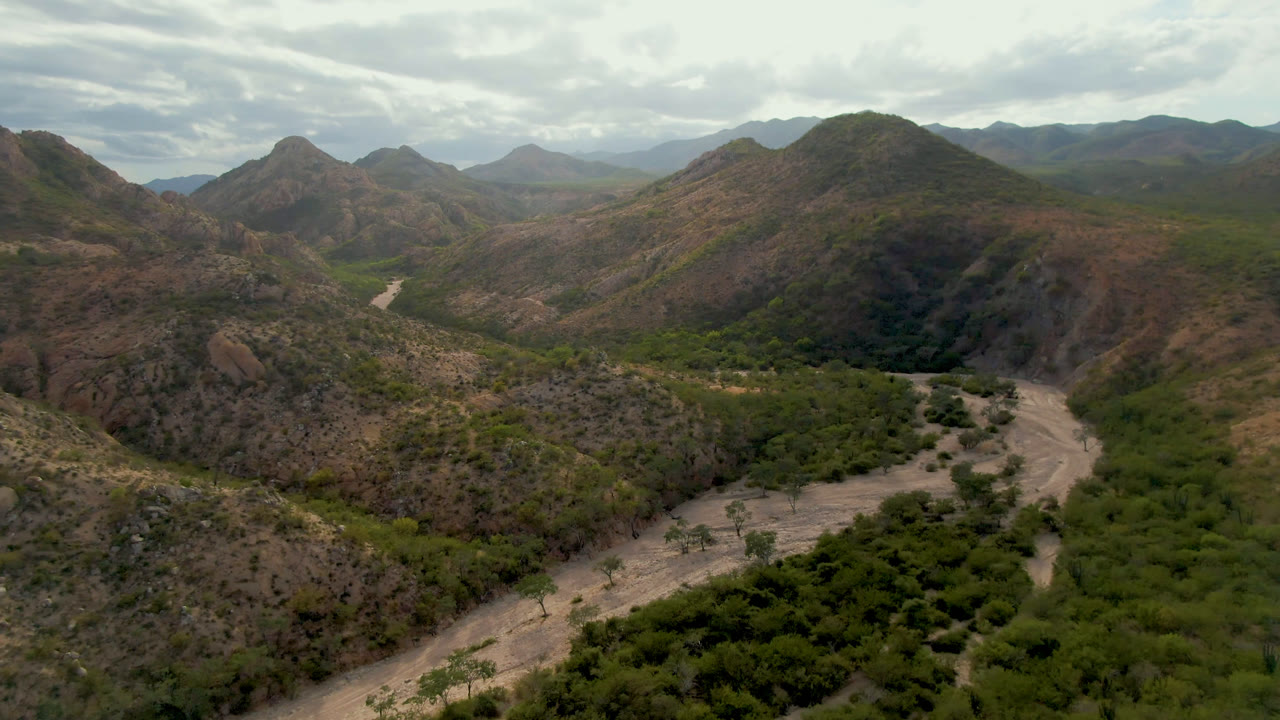 Aerial mountain valley with green vegetation, a pale road, and cloudy daylight.