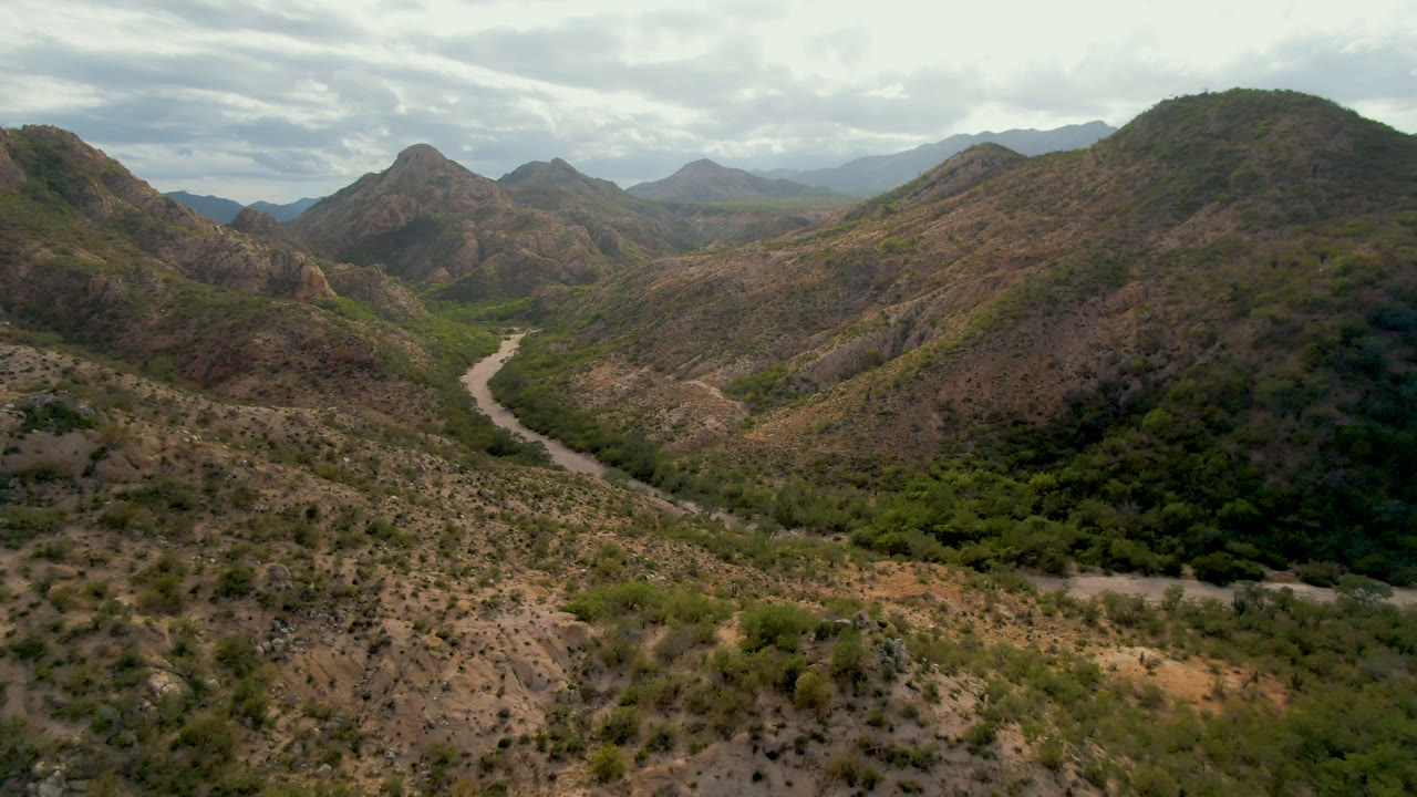 Opening aerial frame over Cabo Pulmo, Baja California Sur, with a pale road curving through dry hills, a green valley floor, distant mountains, cloudy daylight, and no visible people.