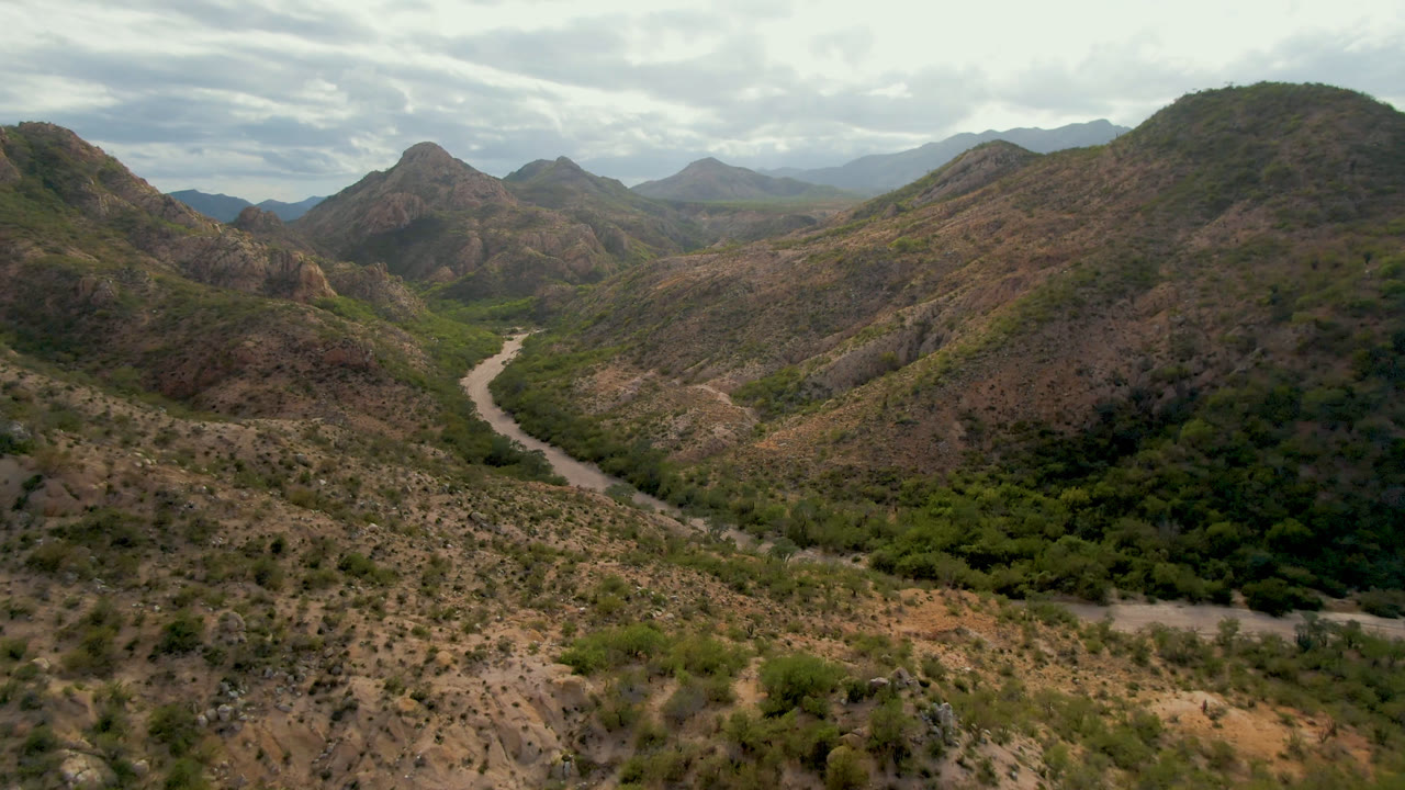 Aerial frame in Cabo Pulmo showing the dirt road more centered between scrubby brown slopes, dense green vegetation below, layered mountain ridges, overcast sky, and no visible people.