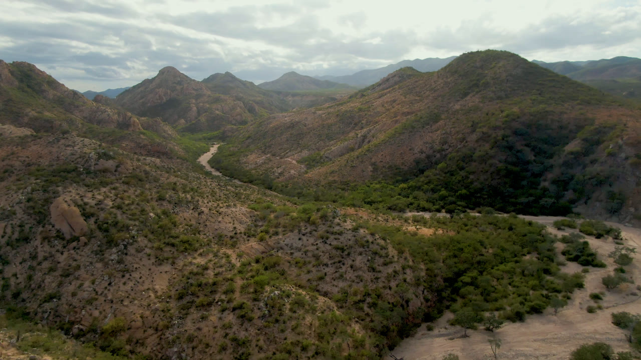 Cabo Pulmo aerial frame with the dirt road running along the left side of a vegetated valley floor, rocky slopes in front, layered hills beyond, and no visible people.