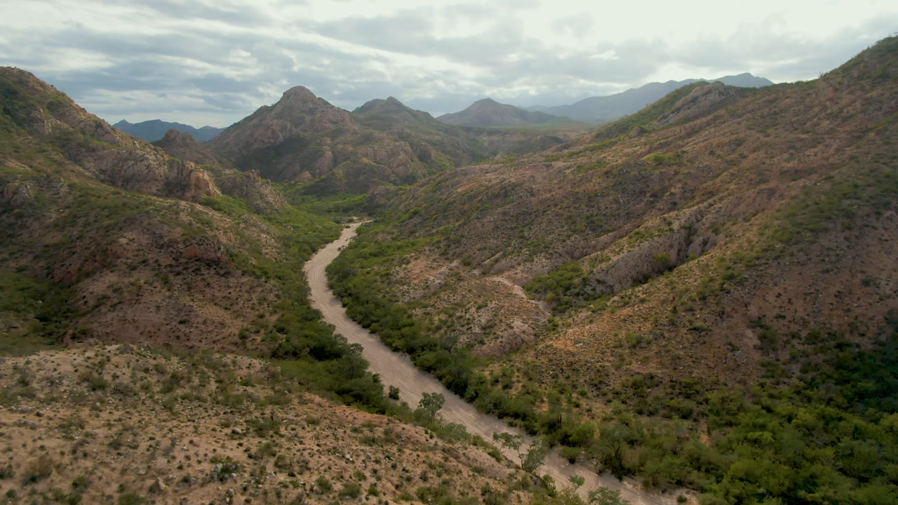 Aerial frame from Cabo Pulmo, Baja California Sur showing a pale winding riverbed or road corridor through green valley vegetation and brown mountain slopes, with no visible people.