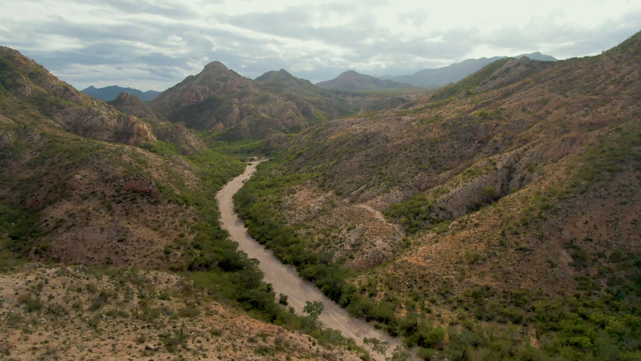 Wide aerial view in Cabo Pulmo with the pale winding corridor shifting closer to center, green vegetation along the valley floor, layered rocky hills, and no visible people.
