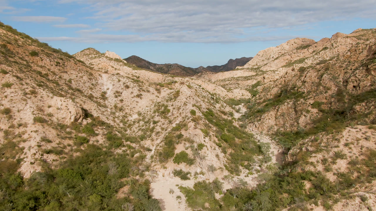 Opening frame of rocky desert hills in Cabo Pulmo, Baja California Sur, with sparse green shrubs, distant dark ridges, thin clouds, and no visible people.