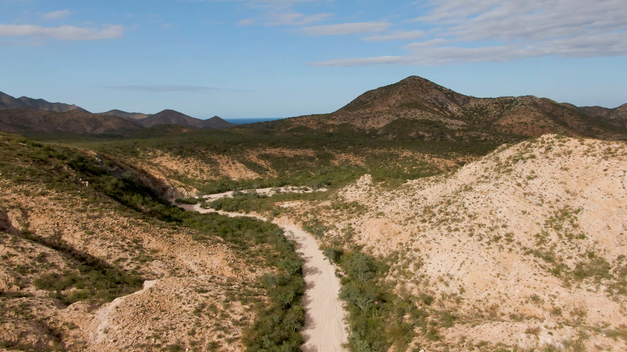 Opening frame of a pale dirt road winding through scrub-covered hills near Cabo Pulmo, Baja California Sur, with distant mountains, blue sky, and no visible people.