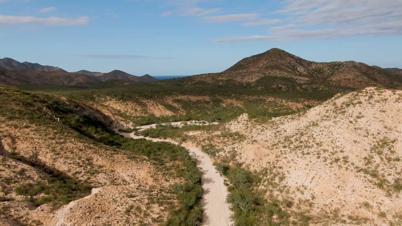 A wide aerial view shows the dirt road crossing the lower valley near Cabo Pulmo, with a rounded mountain centered beyond scrubland and no visible people.