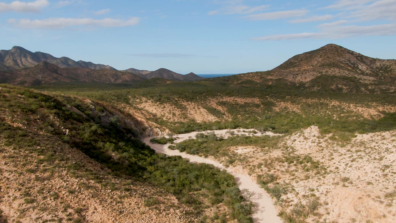 Cabo Pulmo desert frame showing the dirt road lower in the valley, darker brush along the wash, layered mountain ridges, and no visible people.