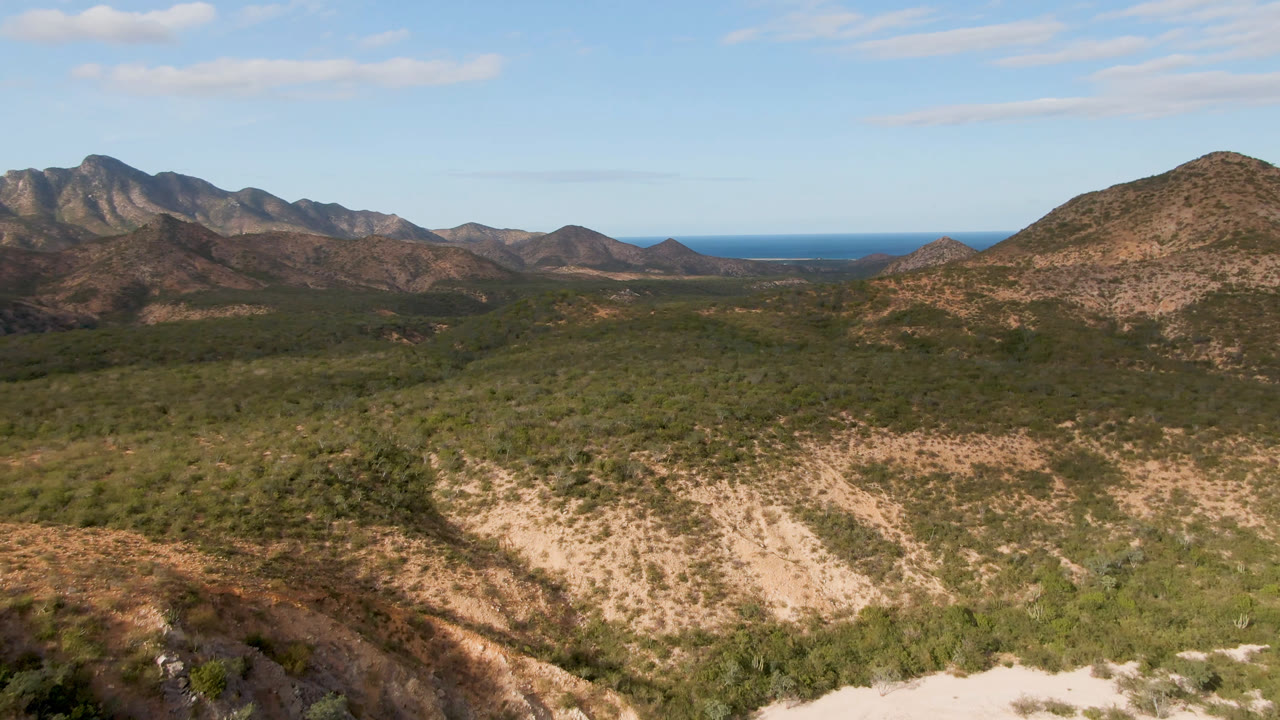 Cabo Pulmo, Baja California Sur landscape with scrub-covered foreground slopes, rugged mountains, distant blue water on the horizon, and no visible people at the opening frame.