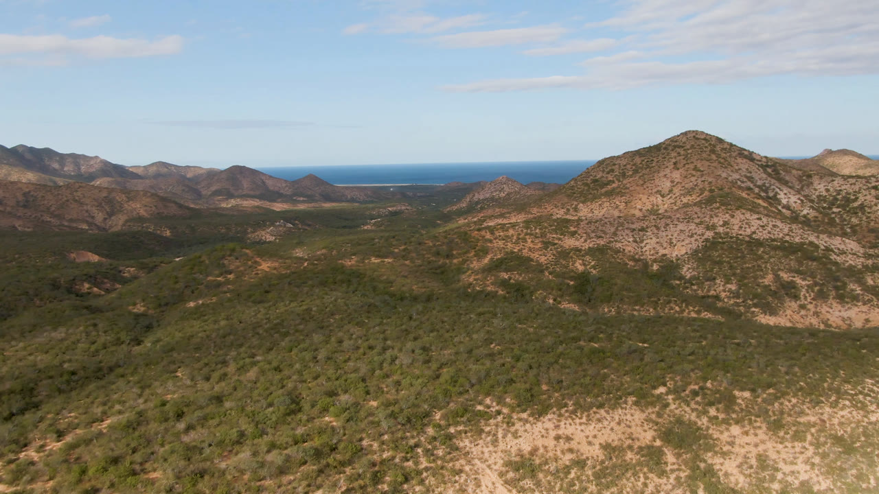 Wide aerial frame from Cabo Pulmo showing a central rocky hill, green-brown desert vegetation, layered ridges, and the ocean line behind them, with no visible people.