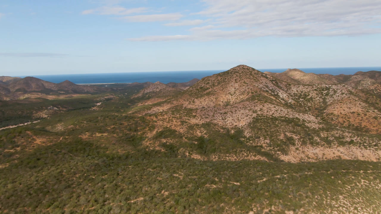 Opening aerial frame over Cabo Pulmo arid mountains, with scrub-covered ridges in the foreground, blue ocean on the horizon, thin cloud cover above, and no visible people.