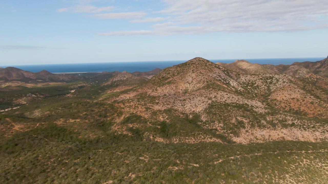 Aerial view in Cabo Pulmo with a central tan ridge, darker green valley vegetation below, distant blue water across the horizon, and no visible people.