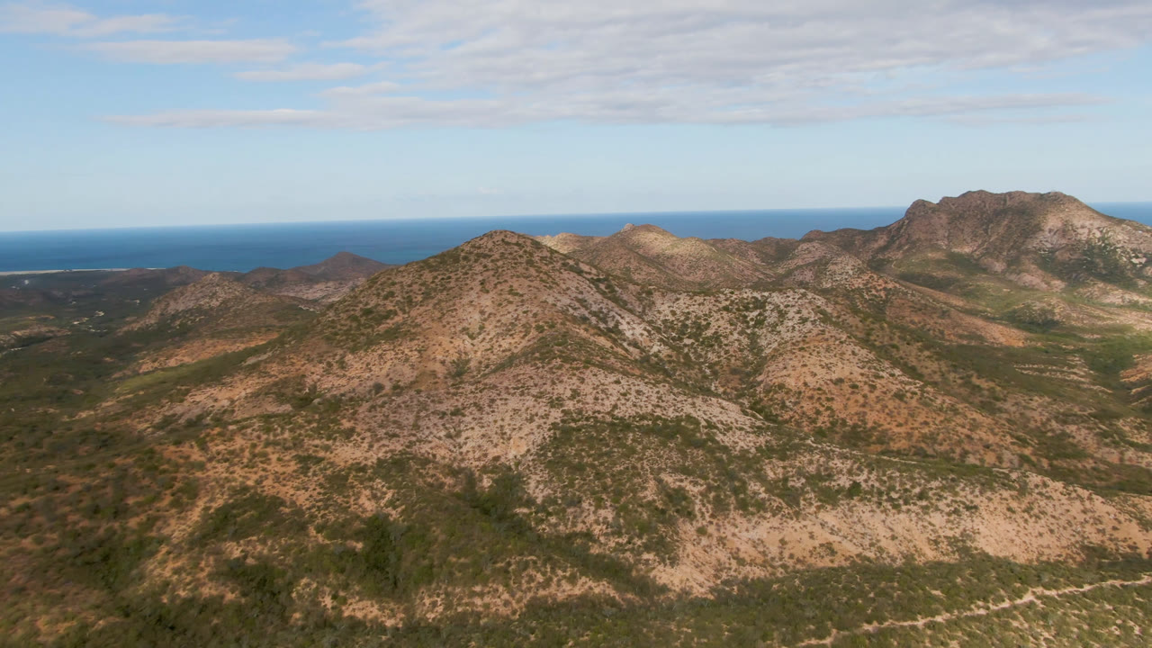 Opening aerial frame over Cabo Pulmo arid mountain ridges with the blue ocean on the horizon, thin cloud cover, and no visible people.