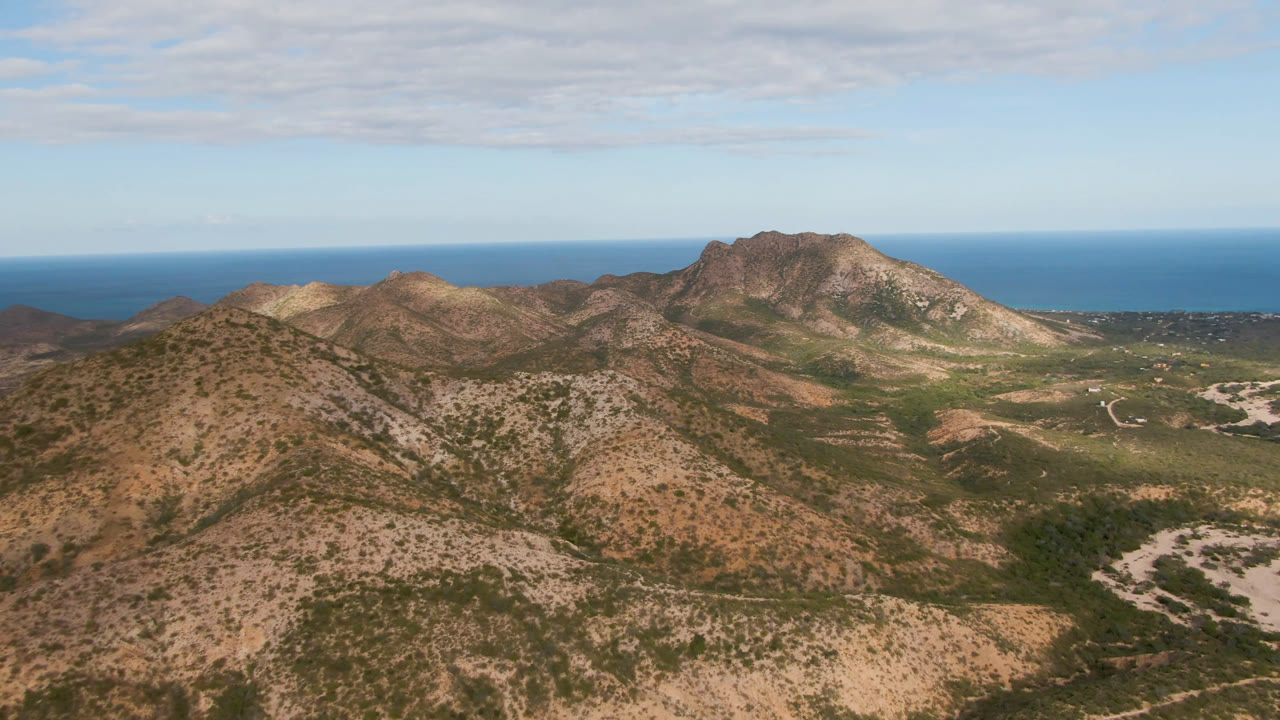 High aerial view over Cabo Pulmo, Baja California Sur showing scrubby tan hills, rocky mountain ridges, blue ocean on the horizon, and no visible people.