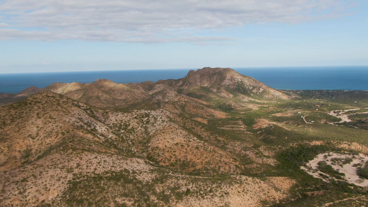 Aerial frame over Cabo Pulmo, Baja California Sur with a rounded scrub-covered hill in front, a pale dry wash across the valley, mountains behind it, ocean at the horizon, and no visible people.