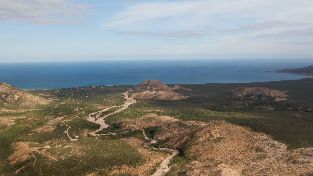 Aerial frame over Cabo Pulmo, Baja California Sur, showing rocky foreground hills, green scrubland, a pale winding dirt road, blue ocean, and no visible people.