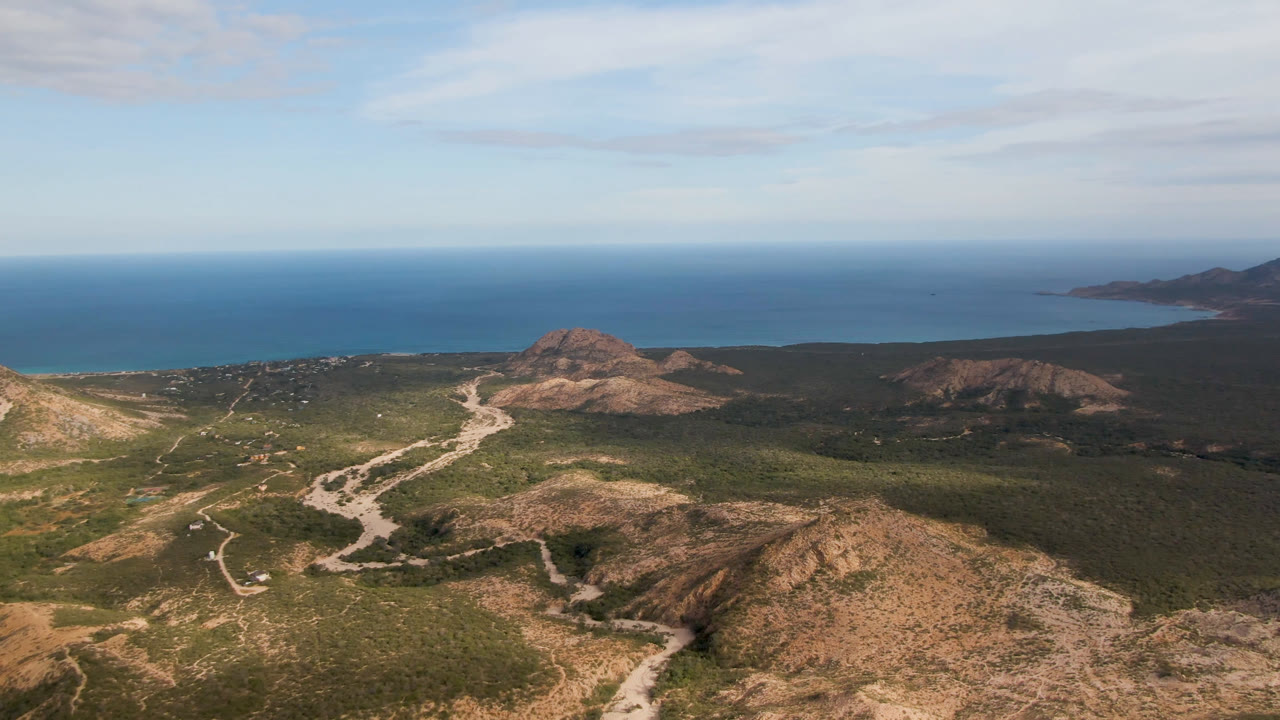 Aerial view in Cabo Pulmo with the winding road more centered through low green terrain, tan hills in front, ocean across the background, and no visible people.