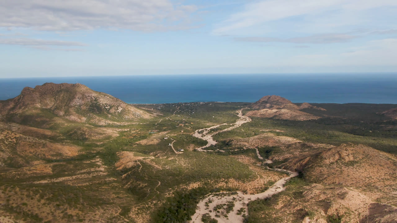 Aerial frame over Cabo Pulmo, Baja California Sur, showing rocky hills, branching dirt roads, green scrub, blue ocean beyond the mountains, and no visible people.