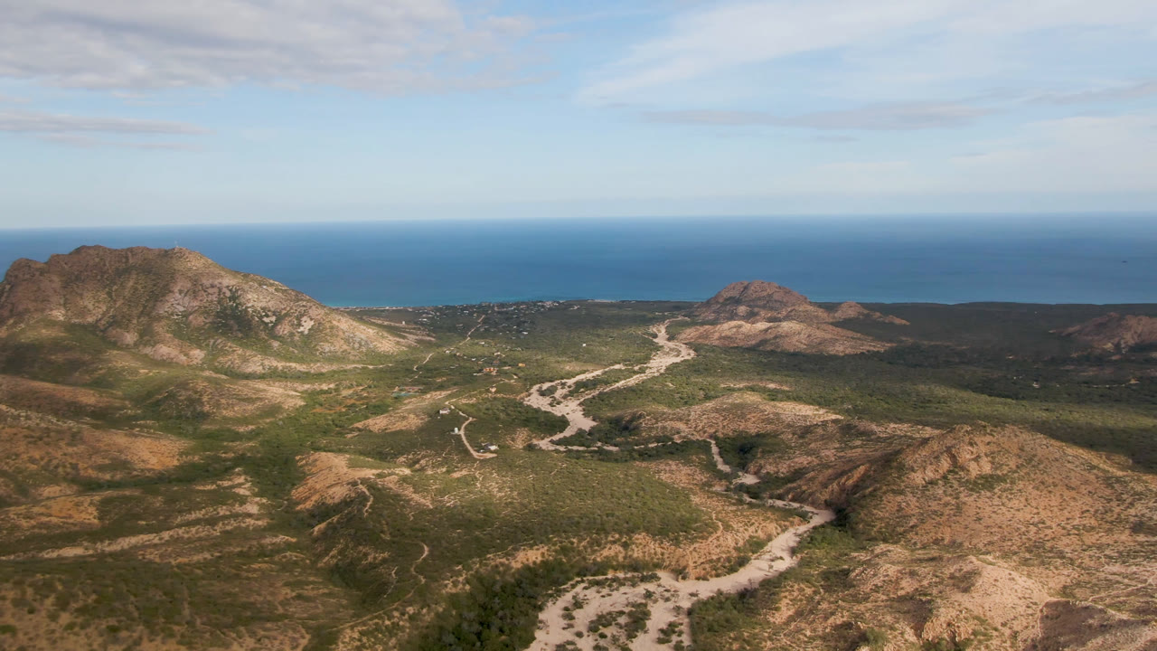 Aerial view in Cabo Pulmo with a large rocky hill on the left, a pale dirt road running through the center valley, scrub-covered slopes, ocean background, and no visible people.