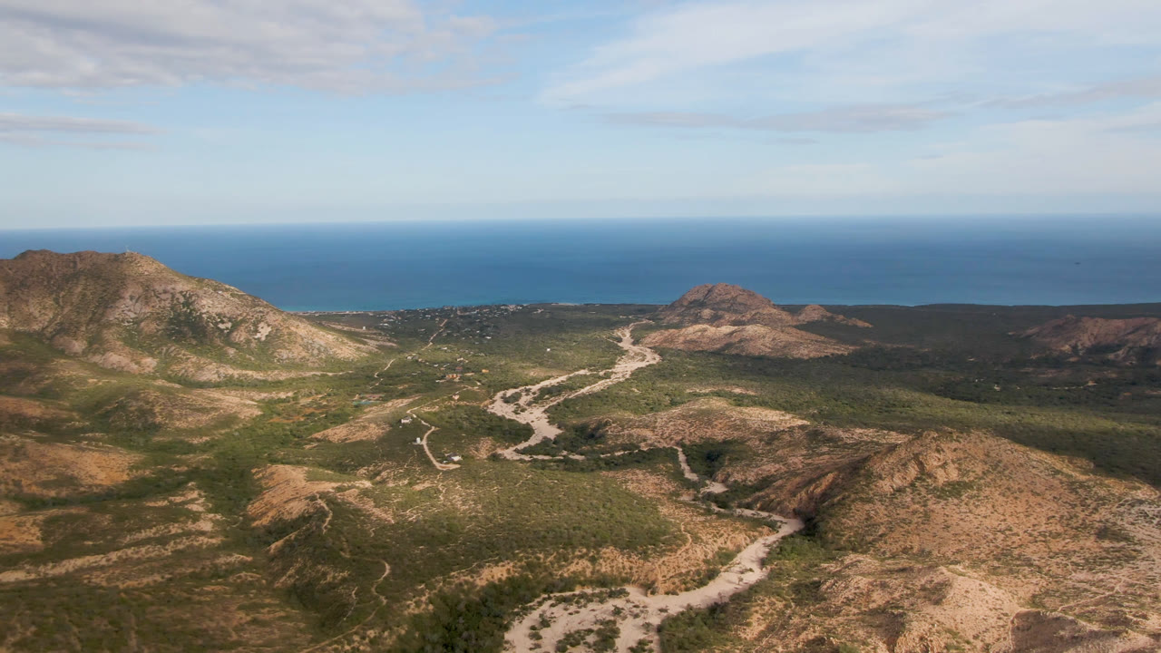Drone perspective over a winding road, low mountains, and blue sea.