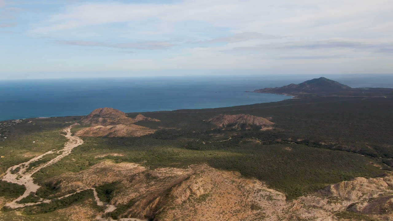 Wide aerial view of rocky hills, scrubland, winding dirt road, and ocean horizon