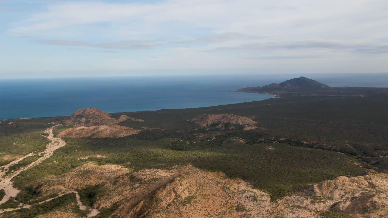 Cabo Pulmo aerial view one second in, with the winding dirt road more centered between rocky hills and dark scrubland leading toward the ocean, with no visible people.