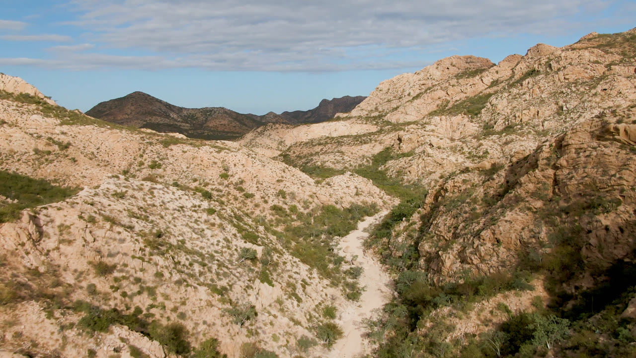 Elevated Cabo Pulmo landscape frame with the dirt road visible through the center valley, tan rock formations on both sides, sparse vegetation, and no visible people.