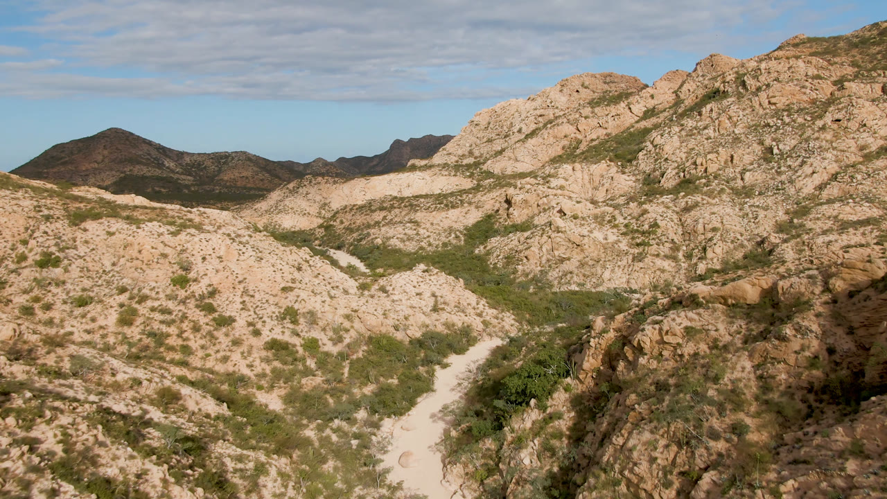 Wide aerial-looking view of rocky hills in Cabo Pulmo, Baja California Sur, with a pale wash cutting through green desert vegetation and no visible people.
