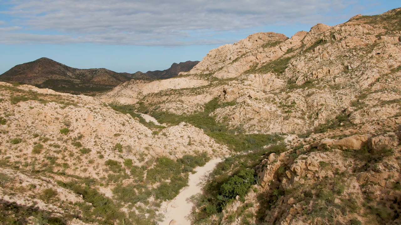 Wide view of Cabo Pulmo rocky slopes with tan ridges, green scrub following the valley floor, distant mountains, and no visible people.