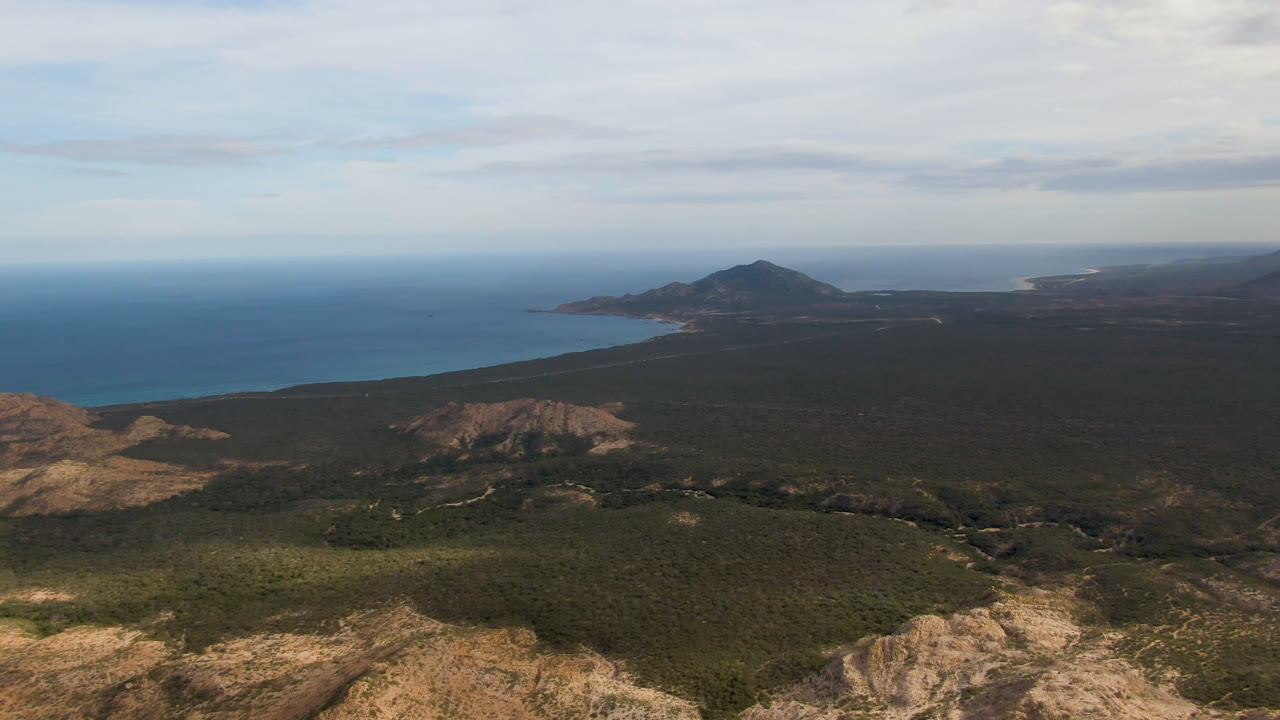 Aerial view over Cabo Pulmo, Baja California Sur, with rocky foreground hills, dark scrubland, blue ocean, distant shoreline, clouded sky, and no visible people.