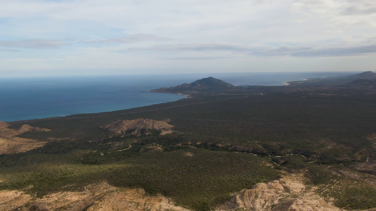 High aerial frame in Cabo Pulmo showing a rocky foreground ridge, broad green-brown scrub plain, blue water to the left, hazy shoreline beyond, and no visible people.